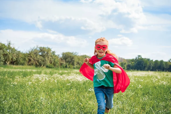 Heureux enfant en rouge super-héros cape et masque courir dans la prairie le jour de l'été — Photo de stock