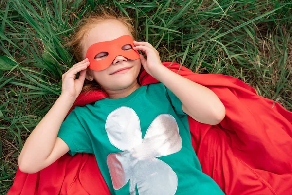 Vista aerea del bambino con gli occhi chiusi in mantello supereroe rosso e maschera sdraiato su erba verde — Foto stock