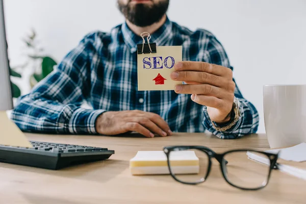 Cropped shot of young bearded developer holding sticker with SEO sign — Stock Photo