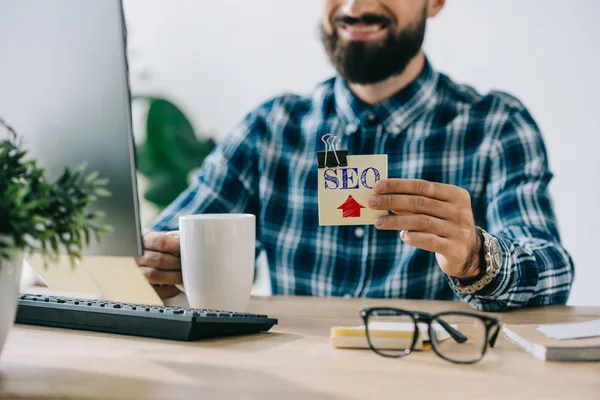 Cropped shot of young smiling bearded developer holding sticker with SEO sign — Stock Photo