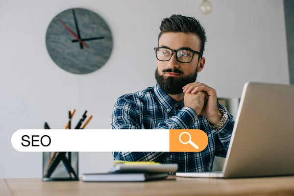 Thoughtful young developer looking away while sitting at workplace with laptop and SEO search — Stock Photo