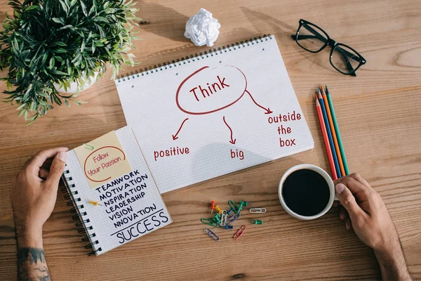 Cropped view of tattooed man with cup of coffee and notebooks with success and think signs — Stock Photo