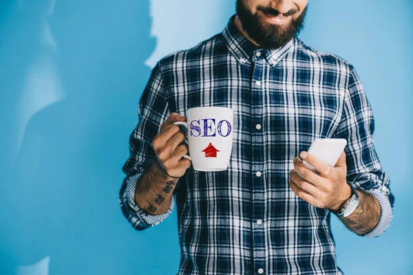 Vue recadrée de l'homme en chemise à carreaux à l'aide d'un smartphone et tenant tasse avec signe SEO — Photo de stock