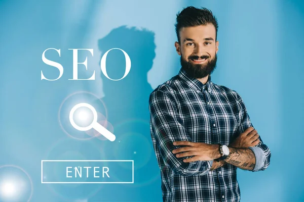Успешный бородатый разработчик позирует со скрещенными руками, на синий с поиском SEO — стоковое фото