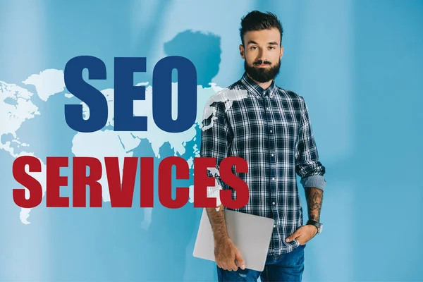 Bearded developer in checkered shirt holding laptop, on blue with world map and SEO services — Stock Photo