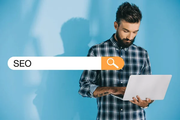 Bearded developer using laptop, on blue with SEO search bar — Stock Photo