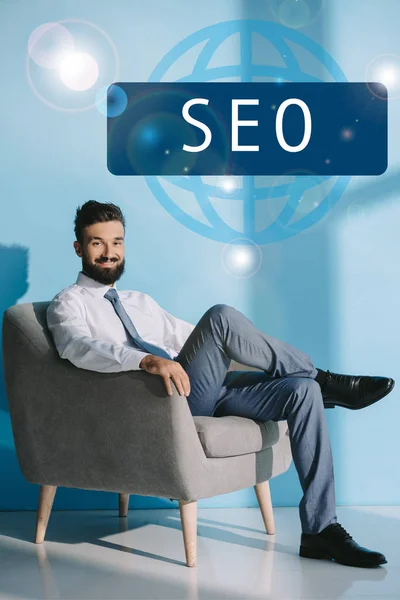 Успішний розробник в офіційному одязі, сидячи в сірому кріслі, на синьому з вивіскою SEO — стокове фото