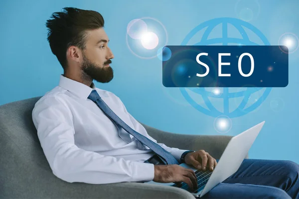 Bearded developer using laptop and sitting in armchair, on blue with SEO sign — Stock Photo