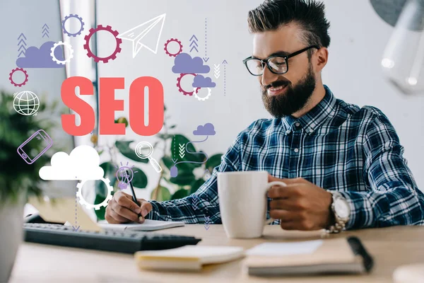 Successful seo manager with cup of coffee and computer writing notes at workplace with SEO icons — Stock Photo