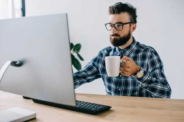 Обрезанный снимок молодого мужчины с чашкой кофе, работающего с компьютером в офисе — стоковое фото