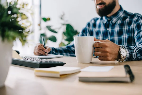 Colpo ritagliato di successo sorridente seo con una tazza di caffè e note di scrittura del computer sul posto di lavoro — Foto stock