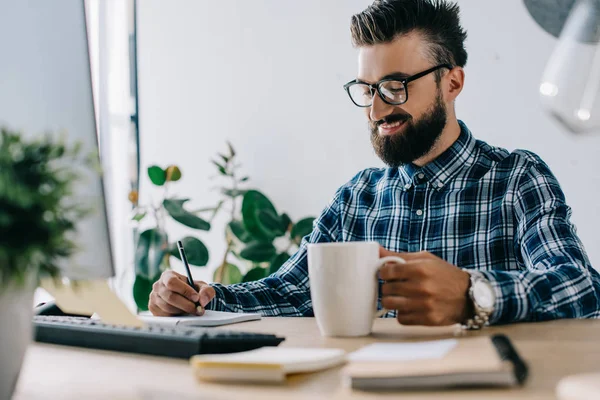 Erfolgreicher lächelnder SEO mit Tasse Kaffee und Computer-Notizen am Arbeitsplatz — Stockfoto