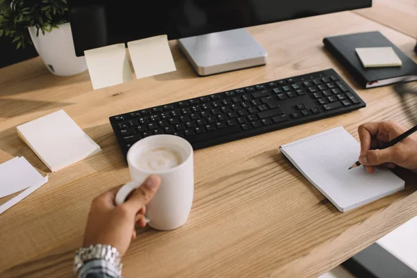 Cropped shot of seo with cup of coffee and computer at workplace writing notes — Stock Photo