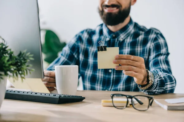 Recortado disparo de joven sonriente barbudo sosteniendo pila de notas en el lugar de trabajo - foto de stock