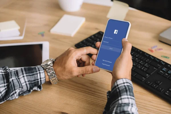 Recortado disparo de hombre usando teléfono inteligente con aplicación de facebook en la pantalla en el lugar de trabajo - foto de stock