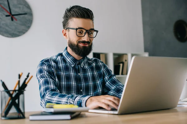 Sorrindo jovem desenvolvedor barbudo trabalhando com laptop — Fotografia de Stock