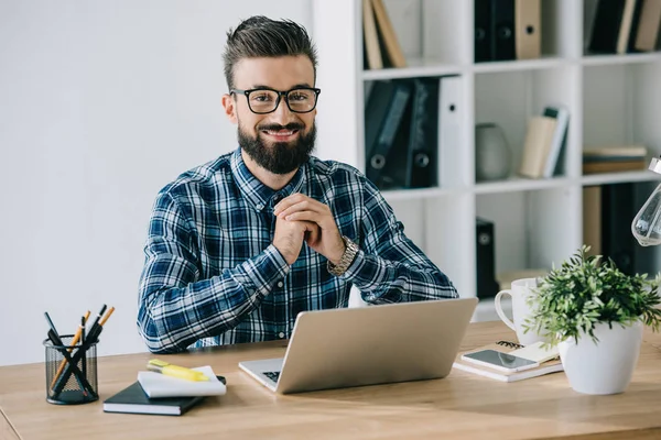 Bearded smiling businessman in eyeglasses looking at camera while sitting at workplace with laptop — Stock Photo