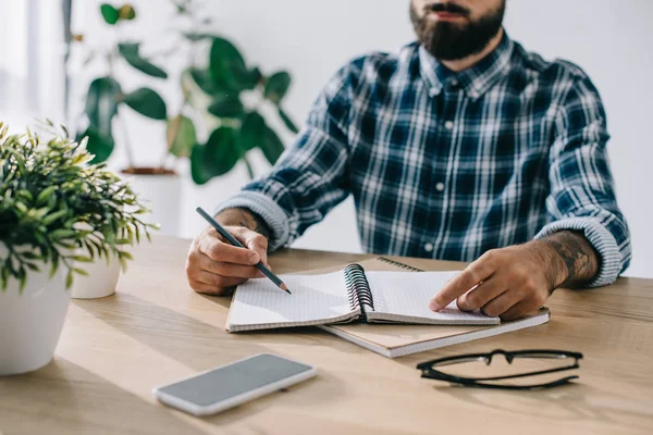 Recortado tiro de hombre en camisa a cuadros escritura en cuaderno en el lugar de trabajo - foto de stock