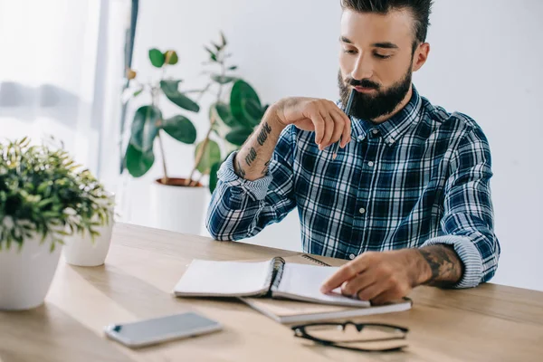 Bel homme réfléchi en chemise à carreaux regardant le carnet — Photo de stock