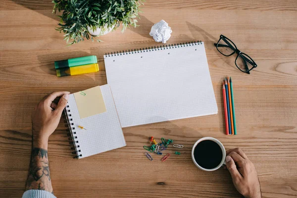 Vista recortada del hombre tatuado con cuadernos y taza de café en el lugar de trabajo - foto de stock