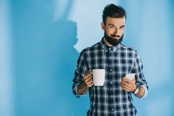 Hombre barbudo guapo con camisa a cuadros sosteniendo teléfono inteligente y taza de café - foto de stock