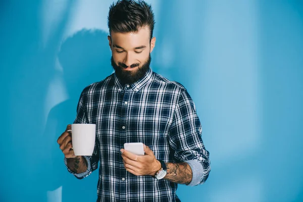 Homme barbu en chemise à carreaux tenant smartphone et tasse de café, sur bleu — Photo de stock