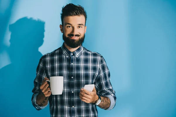 Smiling man in checkered shirt holding smartphone and cup of coffee — Stock Photo