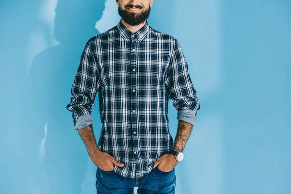 Обрізаний вид бородатого чоловіка, що стоїть з руками в кишенях, на синьому — стокове фото