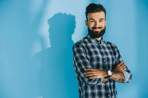 Bärtiger lächelnder Mann, der mit verschränkten Armen posiert und in die Kamera schaut, auf blau — Stockfoto