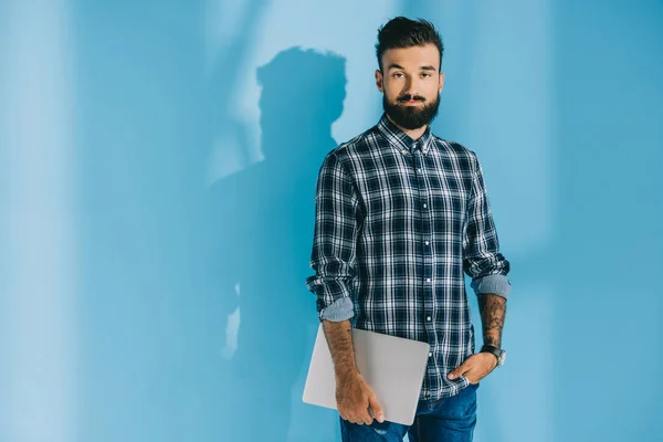 Bearded man in checkered shirt holding laptop, on blue — Stock Photo