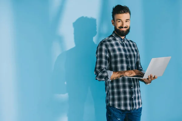 Bel homme souriant en chemise à carreaux utilisant un ordinateur portable, sur bleu — Photo de stock