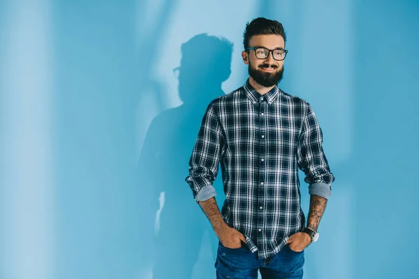 Красивый бородатый мужчина в клетчатой рубашке, стоящий с руками в карманах, на голубой — стоковое фото