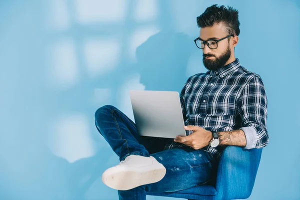 Bearded man in checkered shirt using laptop and sitting in armchair, on blue — Stock Photo