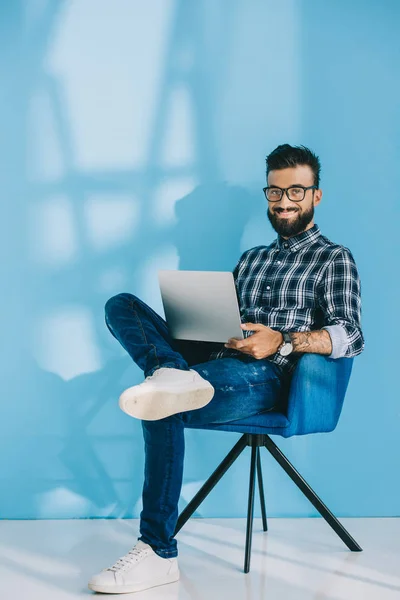 Homme souriant utilisant un ordinateur portable et assis dans un fauteuil, sur bleu — Photo de stock