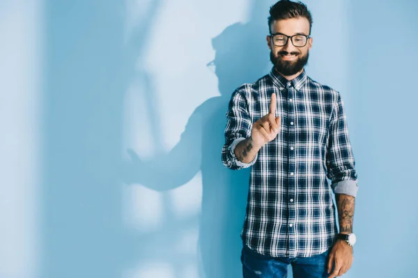 Hombre barbudo sonriente con camisa a cuadros apuntando a algo, en azul - foto de stock