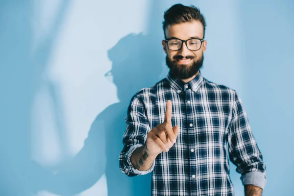 Hombre sonriente con anteojos y camisa a cuadros apuntando a algo, en azul - foto de stock
