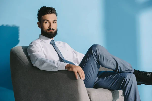 Hombre de negocios guapo en ropa formal sentado en sillón, en azul - foto de stock