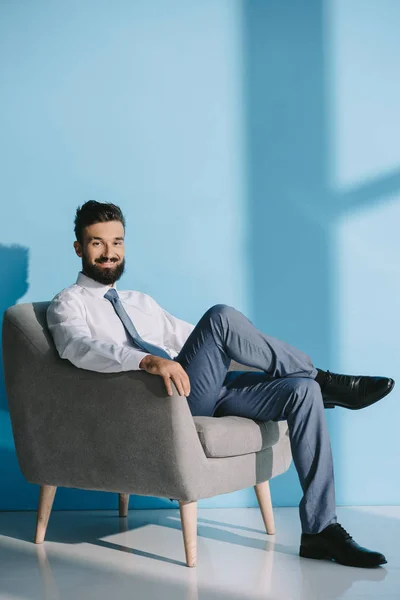 Homme d'affaires souriant en tenue de cérémonie assis dans un fauteuil gris, sur bleu — Photo de stock