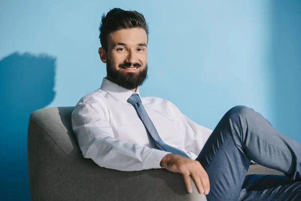 Barbudo sonriente hombre de negocios en ropa formal sentado en sillón, en azul - foto de stock