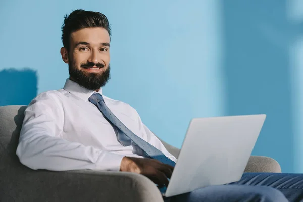 Handsome smiling businessman in formal wear using laptop, on blue — Stock Photo