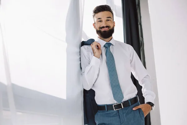 Homme d'affaires souriant tenant veste et debout près de la fenêtre dans le bureau — Photo de stock
