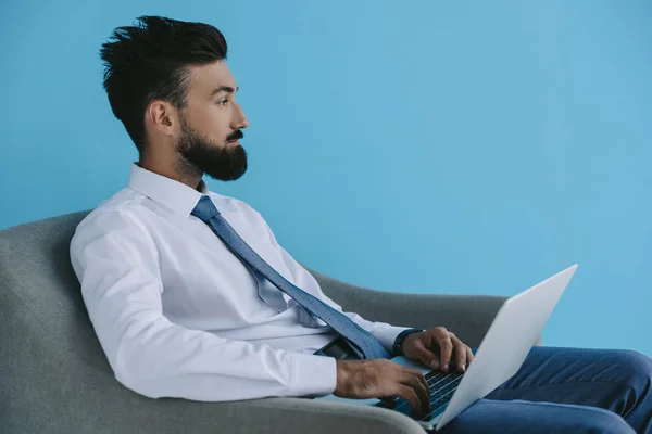 Bearded businessman using laptop and sitting in armchair, on blue — Stock Photo