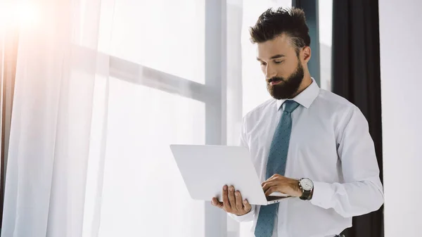 Bell'uomo d'affari barbuto che utilizza il computer portatile vicino alla finestra in ufficio — Foto stock