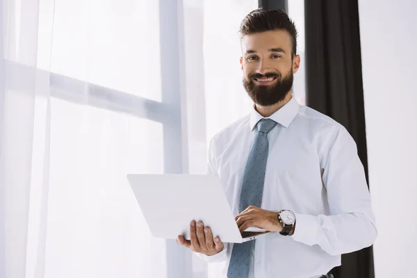 Homem de negócios sorridente barbudo usando laptop perto da janela no escritório — Fotografia de Stock