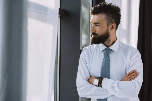 Homme d'affaires barbu avec les bras croisés regardant la fenêtre dans le bureau — Photo de stock