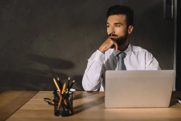 Стомлений бородатий бізнесмен сидить на робочому місці з ноутбуком і олівцями — стокове фото