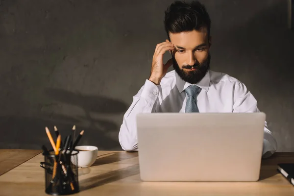 Hombre de negocios reflexivo utilizando el ordenador portátil en el lugar de trabajo con café y lápices - foto de stock