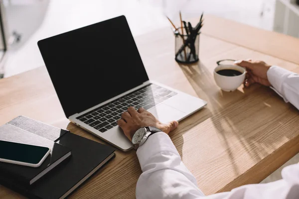 Cropped view of businessman using laptop with blank screen and holding cup of coffee — Stock Photo