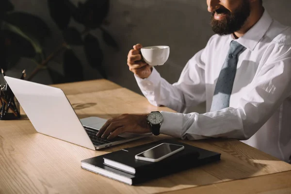 Vista ritagliata di uomo d'affari utilizzando il computer portatile e tenendo in mano una tazza di caffè sul posto di lavoro — Stock Photo