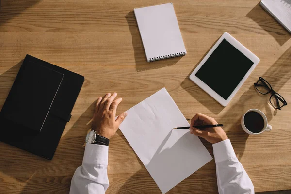 Ausgeschnittene Ansicht eines Mannes, der am Arbeitsplatz mit Kaffee und digitalem Tablet auf Papier schreibt — Stockfoto